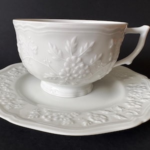 May include: A white porcelain teacup and matching saucer set featuring a raised, embossed floral pattern. The teacup has a curved handle and a scalloped rim, while the saucer displays a matching embossed botanical design around its edge.