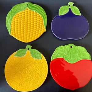 May include: Four ceramic trivets in the shapes of produce. The trivets are a yellow corn, a purple eggplant, a yellow lemon, and a red tomato. Each trivet has a green leaf design at the top and a hole for hanging.