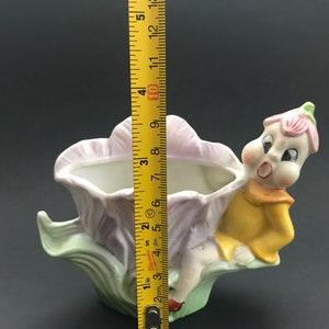 May include: A ceramic planter shaped like a flower with a whimsical fairy figure sitting on the side. The planter is painted in shades of pink, green, and yellow.