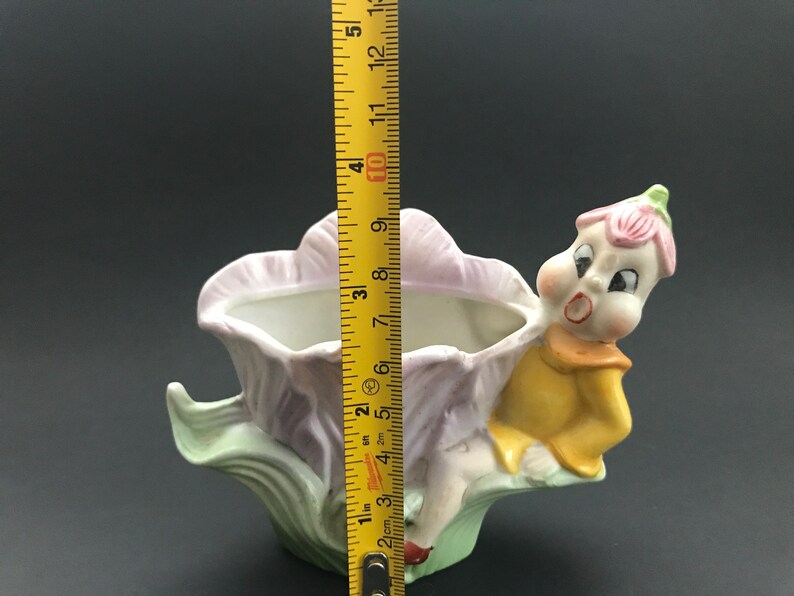 May include: A ceramic planter shaped like a flower with a whimsical fairy figure sitting on the side. The planter is painted in shades of pink, green, and yellow.