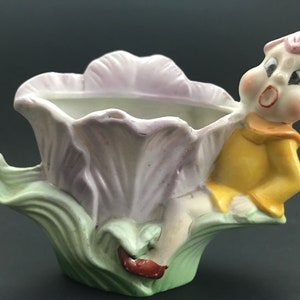 May include: A ceramic planter shaped like a flower with a small elf figurine sitting on the side. The flower is purple and green, and the elf is wearing a yellow shirt and red shoes.
