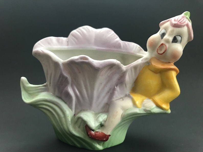 May include: A ceramic planter shaped like a flower with a small elf figurine sitting on the side. The flower is purple and green, and the elf is wearing a yellow shirt and red shoes.