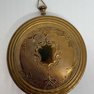 May include: A round, gold-toned metal compact with an intricate engraved pattern featuring a central shield-shaped cartouche surrounded by cross-hatching and leaf motifs. It includes a small, triangular hanging loop at the top of the casing.