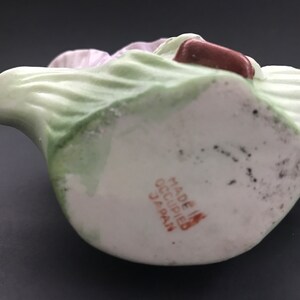 May include: A white porcelain figurine of a woman in a green dress and a pink hat. The figurine is holding a book and has a brown base with the text "Made in Occupied Japan" printed on it.
