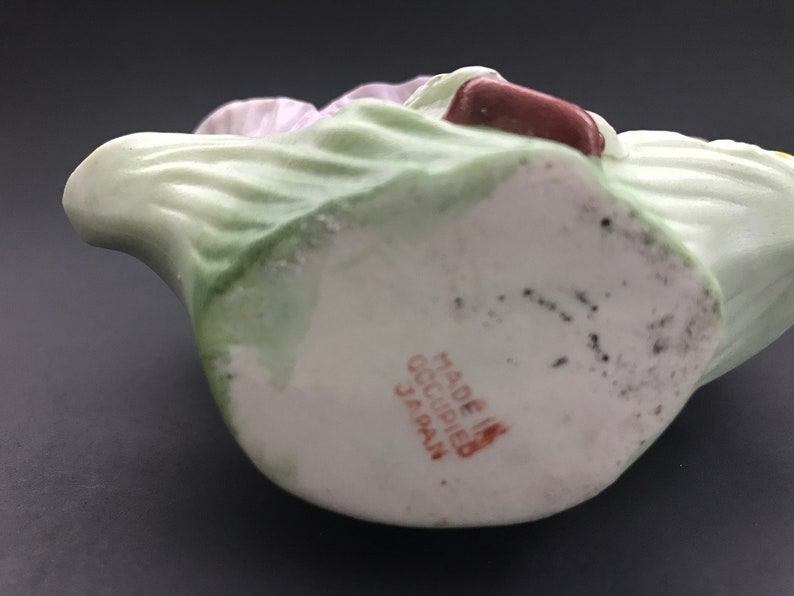 May include: A white porcelain figurine of a woman in a green dress and a pink hat. The figurine is holding a book and has a brown base with the text "Made in Occupied Japan" printed on it.