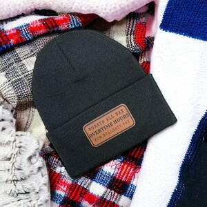 May include: A black beanie with a brown rectangular patch. The patch has white text that reads "WORKIN ALL DAY OVERTIME HOURS FOR BULLSHIT PAY". The beanie is resting on a plaid and striped fabric.