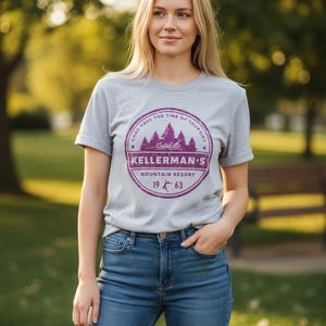 Op de afbeelding: Lichtgrijs t-shirt met een paarse cirkelvormige afbeelding met de tekst "Kellerman's Mountain Resort 1963". De persoon draagt een blauwe spijkerbroek. De achtergrond is een vage buitenscène met bomen.