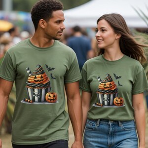 May include: Two people wearing olive green t-shirts with a Halloween cupcake graphic. The cupcake has orange frosting, a black and white striped wrapper, and a jack-o'-lantern face. Bats and a pumpkin are also featured.