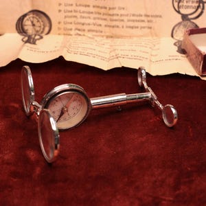Puede incluir: Una lupa vintage plateada con una esfera de brújula, abierta y apoyada sobre una superficie de terciopelo rojo oscuro. La lupa tiene dos lentes circulares y un mango. Un trozo de papel envejecido con texto y una pequeña caja están en el fondo.