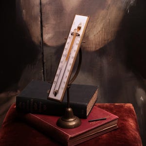 May include: A vintage-style thermometer mounted on a brass stand with a white rectangular face and black numerical markings. The thermometer features a glass tube containing a yellow liquid and is positioned upright on a brass base.