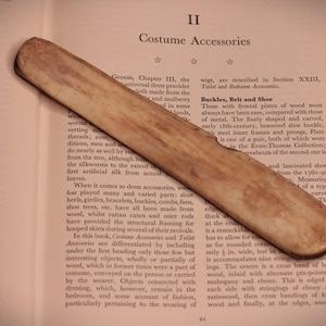 Könnte beinhalten: Ein langes, glattes, helles Holzwerkzeug mit abgerundeter, spitz zulaufender Form, das diagonal auf einer offenen Buchseite liegt. Das Objekt weist eine natürliche, gesprenkelte Maserung mit subtilen braunen und beigen Farbnuancen auf.