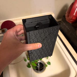 Peut inclure: Un pot noir rectangulaire avec un motif texturé et répétitif. Le pot est tenu au-dessus d'un évier, l'eau s'écoulant du fond. Des feuilles et des vignes vertes sont visibles à l'intérieur du pot.