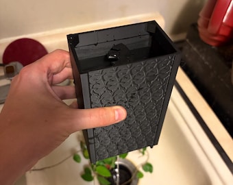 May include: A black rectangular planter with a textured, repeating pattern. The planter is held over a sink, with water draining from the bottom. Green plant leaves are visible inside the planter.