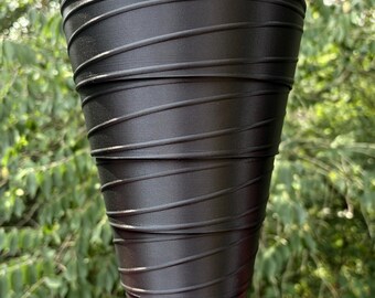 May include: A black, cylindrical planter with a textured, ridged design. The planter has a wide top and tapers down to a smaller base. The ridges are horizontal and wrap around the planter. The background is blurred green foliage.