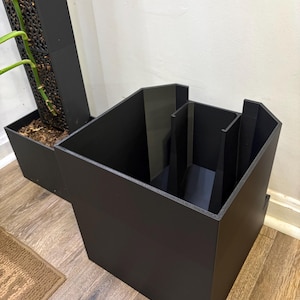 Peut inclure: Un bac à plantes noir géométrique avec une conception à compartiments multiples. Le bac est fait d'un matériau sombre et solide et repose sur un sol en bois. Un deuxième bac à plantes plus grand est visible en arrière-plan.