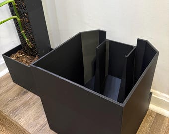 May include: A black, geometric planter box with a multi-compartment design. The planter is made of a solid, dark material and sits on a wooden floor. A second, taller planter is visible in the background.