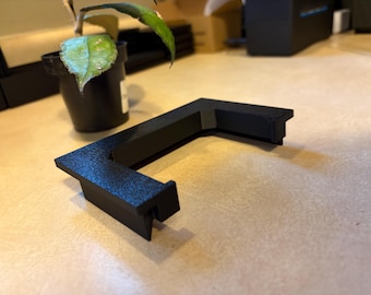 May include: A black, L-shaped bracket with a textured surface. The bracket is made of a solid material and has a notch on one end. A small potted plant is visible in the background.