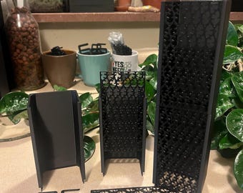 May include: A collection of black metal plant accessories, including a rectangular planter, a decorative screen with a floral pattern, and several small clips. The items are arranged on a light-colored surface, with plants visible in the background.