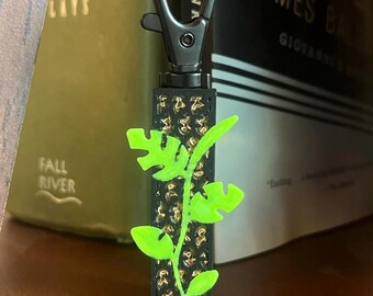 May include: A black keychain with a silver clasp and a rectangular body featuring a gold pattern. A neon green plant design is attached to the body. The keychain is resting on a wooden surface.