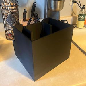 May include: A black plastic organiser with multiple compartments for storing kitchen utensils. The organiser is sitting on a light brown worktop.