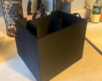 May include: A black plastic organizer with multiple compartments for storing kitchen utensils. The organizer is sitting on a light brown countertop.