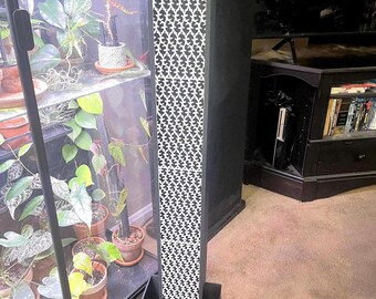 May include: A tall, black, rectangular air purifier with a white patterned front. The purifier is sitting on a black base and is in front of a glass cabinet with plants.