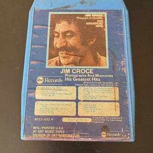Puede incluir: Una caja de cassette azul con una foto de Jim Croce en la parte delantera. La caja tiene el título "Jim Croce Photographs & Memories His Greatest Hits" y el texto "ABC Records" en la parte delantera. La parte posterior de la caja enumera las canciones de la cinta, incluyendo "Bad, Bad Leroy Brown", "Operator (That's Not The Way It Feels)", "I'll Say It To Say I Love You In A Song", y "Got A Name".