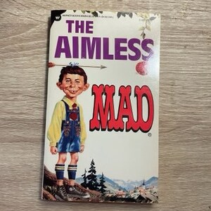 Puede incluir: Una portada de la revista Mad vintage con un personaje de dibujos animados que lleva un corazón rojo y un atuendo azul y blanco. El título de la portada es "The Aimless Mad".