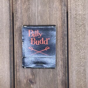 Puede incluir: Una caja de cerillas negra con el texto "Billy Budd" en letras rojas y un par de espadas cruzadas.
