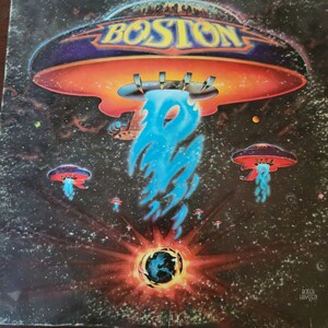 Op de afbeelding: Een vintage vinyl platenhoes met een psychedelische illustratie van een vliegende schotel met het woord "BOSTON" in grote letters. De schotel is omringd door sterren en een gloeiend blauw licht. De platenhoes is in goede staat met wat lichte slijtage.
