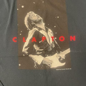 Könnte beinhalten: Schwarzes T-Shirt mit einem weißen Bild von Eric Clapton, der Gitarre spielt. Der Text "CLAPTON" ist in roten Buchstaben über dem Bild gedruckt.