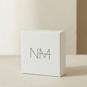 May include: A white, textured box with the logo "NEMO MATHEO" printed in black. The box is rectangular and sits on a neutral-toned surface. The logo features stylized letters and numbers.