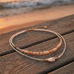 May include: Two layered necklaces with a beachy aesthetic. The top necklace features small, round, white beads and a section of peach-colored beads. The bottom necklace has a large, pearlescent pendant and gold accents. The necklaces rest on a wooden surface.