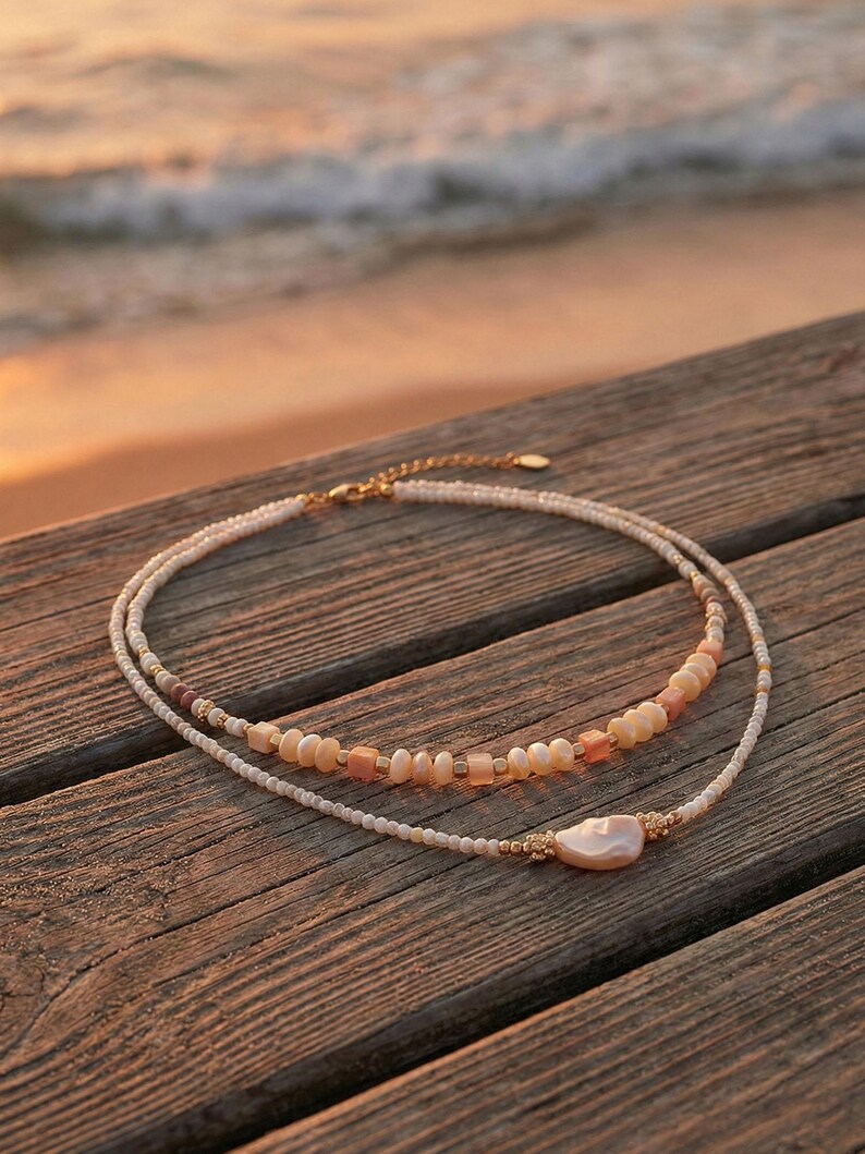 May include: Two layered necklaces with a beachy aesthetic. The top necklace features small, round, white beads and a section of peach-colored beads. The bottom necklace has a large, pearlescent pendant and gold accents. The necklaces rest on a wooden surface.
