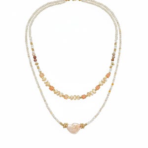 May include: A layered necklace featuring two strands of white beads with gold accents. The top strand includes peach and brown beads, while the bottom strand has a heart-shaped pendant. The necklace is designed for a delicate and feminine look.