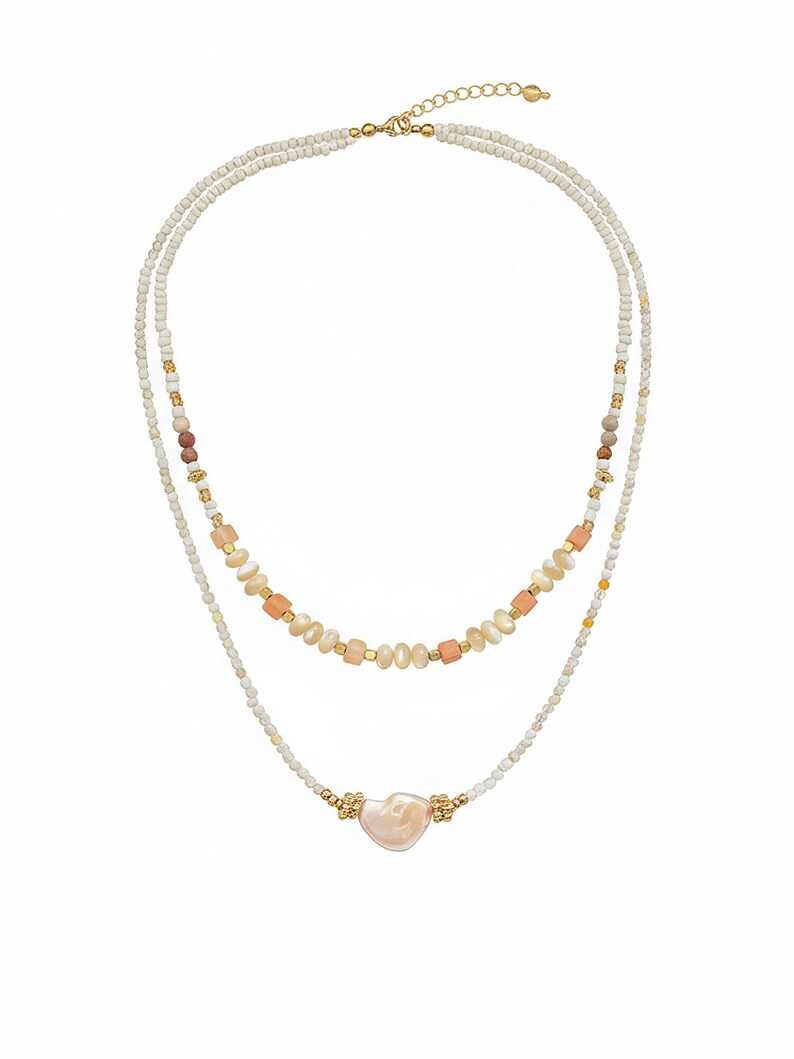 May include: A layered necklace featuring two strands of white beads with gold accents. The top strand includes peach and brown beads, while the bottom strand has a heart-shaped pendant. The necklace is designed for a delicate and feminine look.