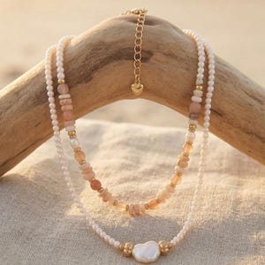 May include: Two layered necklaces with a beachy aesthetic. The top necklace features peach-colored stone beads and gold accents. The bottom necklace has a large, irregular pearl pendant and small white pearls. The necklaces are displayed on a piece of driftwood.