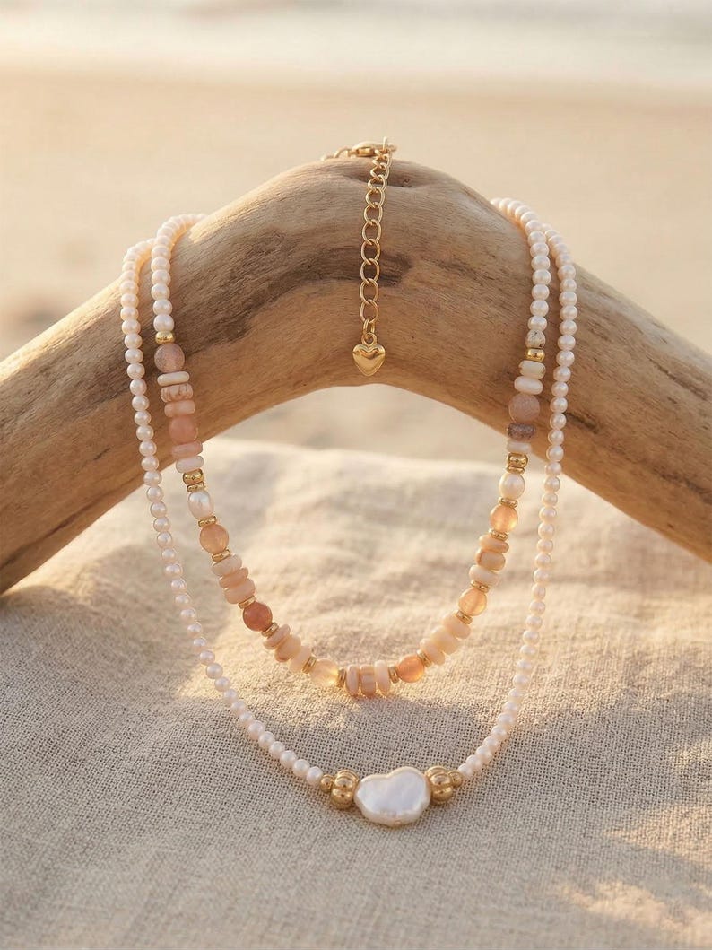 May include: Two layered necklaces with a beachy aesthetic. The top necklace features peach-colored stone beads and gold accents. The bottom necklace has a large, irregular pearl pendant and small white pearls. The necklaces are displayed on a piece of driftwood.