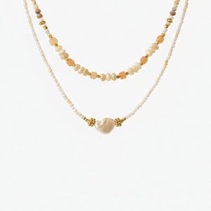 May include: Two layered necklaces. The top necklace features a strand of small white beads, interspersed with peach and gold-colored beads. The bottom necklace has a heart-shaped pearl pendant with gold accents. Both necklaces are on a white background.