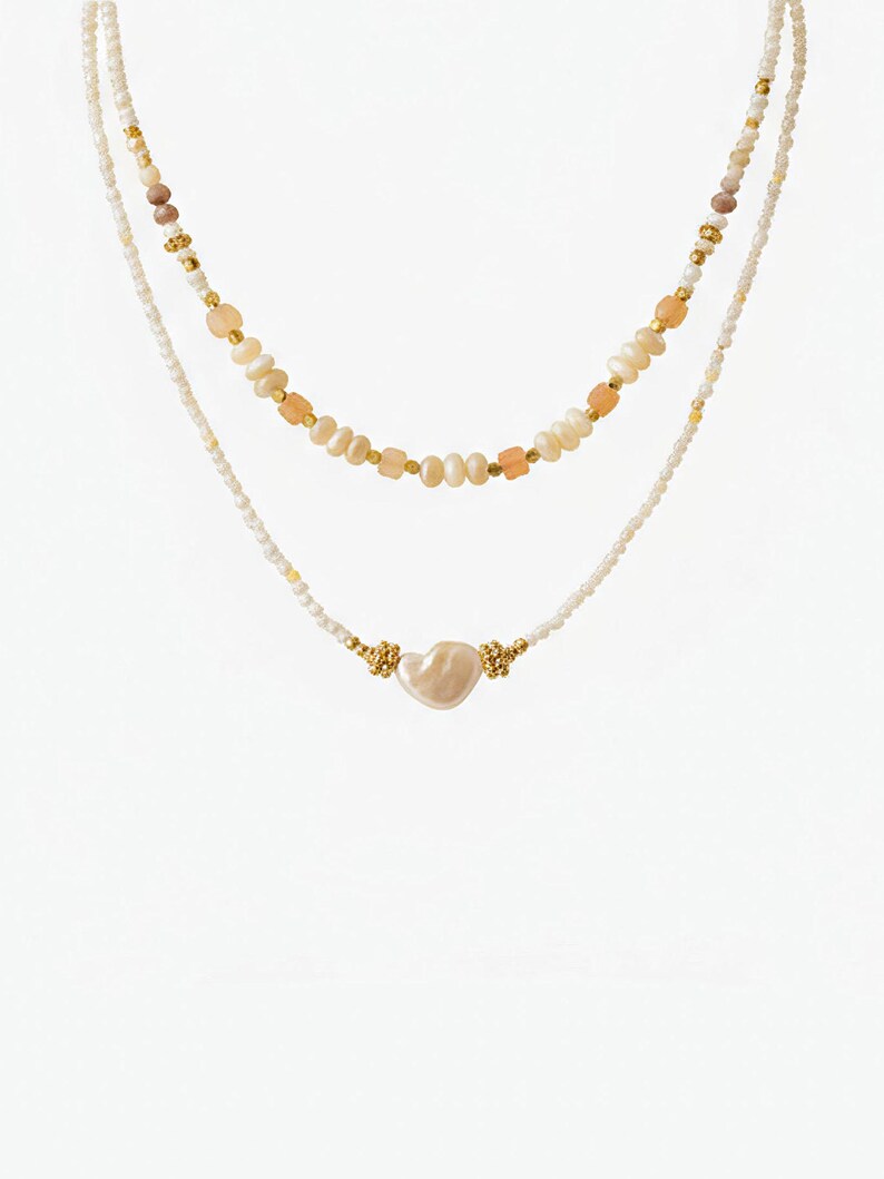 May include: Two layered necklaces. The top necklace features a strand of small white beads, interspersed with peach and gold-colored beads. The bottom necklace has a heart-shaped pearl pendant with gold accents. Both necklaces are on a white background.