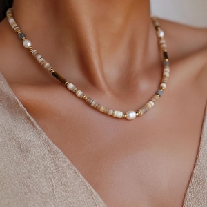 Puede incluir: Un collar de cuentas con una mezcla de cuentas de disco de tonos naturales, pequeñas cuentas doradas y perlas de agua dulce. El collar tiene un diseño delicado y se usa alrededor del cuello.
