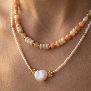 May include: Two layered necklaces. The top necklace features peach and white beads with gold accents. The bottom necklace has small white pearls and a heart-shaped pearl pendant with gold floral details. Jewelry.
