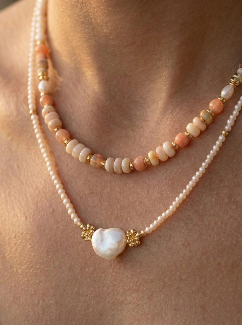 May include: Two layered necklaces. The top necklace features peach and white beads with gold accents. The bottom necklace has small white pearls and a heart-shaped pearl pendant with gold floral details. Jewelry.