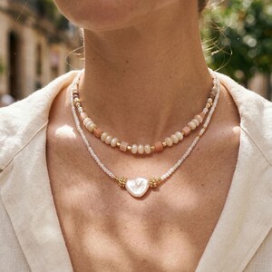 May include: Two layered necklaces. The top necklace features a strand of peach-colored beads and gold accents. The bottom necklace has a heart-shaped pendant with gold details. The necklaces are on a person wearing a cream-colored shirt.