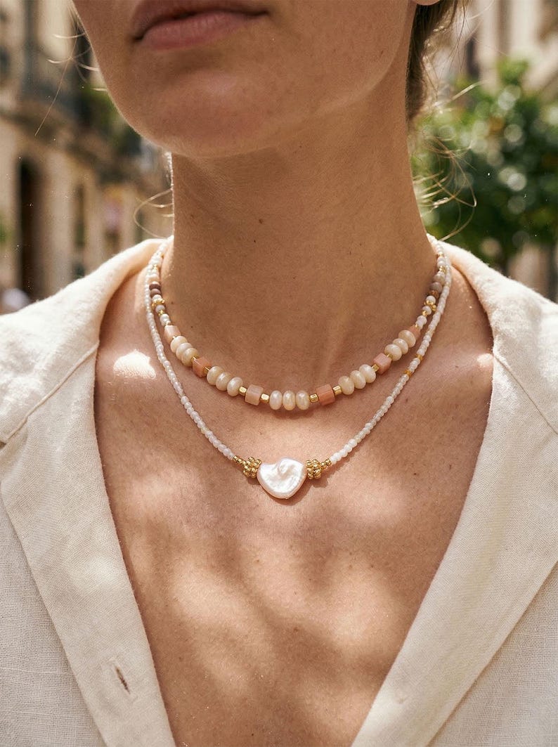 May include: Two layered necklaces. The top necklace features a strand of peach-colored beads and gold accents. The bottom necklace has a heart-shaped pendant with gold details. The necklaces are on a person wearing a cream-colored shirt.