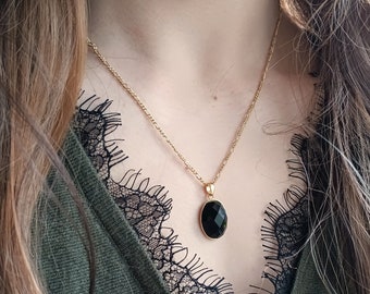 Op de afbeelding: Een gouden ketting met een zwarte ovale hanger. De ketting wordt gedragen door een persoon met lang bruin haar en een zwart kanten topje.