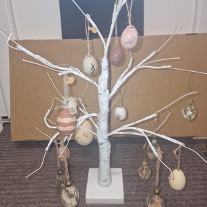 Op de afbeelding: Een witte berkenboom met een witte basis, versierd met verschillende pastelkleurige en heldere glazen paaseieren. De eieren hangen aan de takken van de boom.