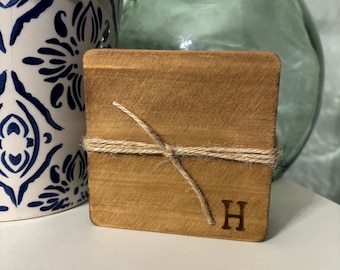 Puede incluir: Un posavasos cuadrado de madera con la letra "H" grabada. El posavasos está atado con hilo. Una maceta estampada azul y blanca con vegetación y un jarrón de cristal verde están en el fondo.