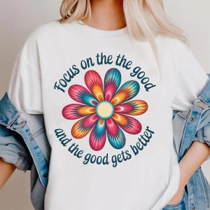 Peut inclure: T-shirt blanc avec un motif floral coloré aux pétales roses, bleus, oranges et jaunes. Le texte "Focus on the good and the good gets better" est imprimé autour de la fleur.