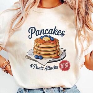 Könnte beinhalten: Cremefarbenes T-Shirt mit einem Stapel Pfannkuchen, bedeckt mit Sirup, Butter und Blaubeeren. Der Text "Pancakes & Panic Attacks" ist in einer verspielten Schriftart dargestellt. Ein "Kaufe eins, erhalte eins"-Grafik ist ebenfalls sichtbar.